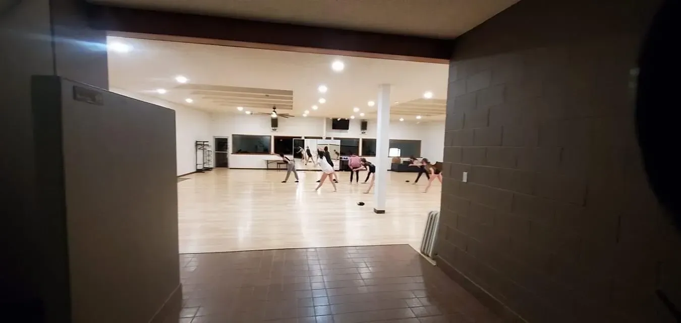 Dancers in a studio, viewed from a doorway. Warm lighting illuminates the wooden floor and people in dance poses.