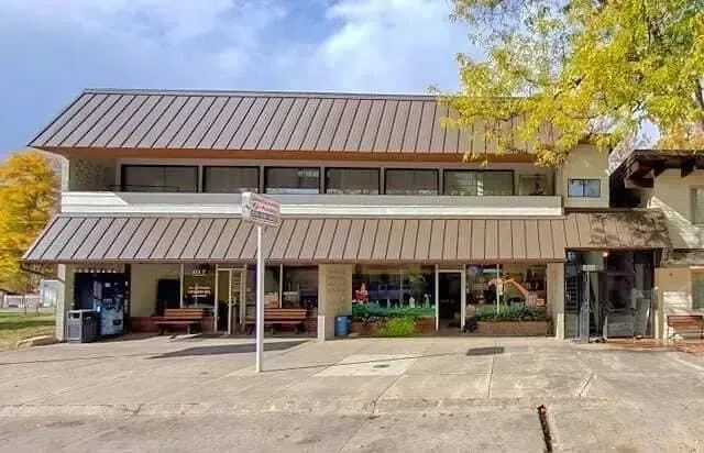 Two-story building with tan roof, awnings, and large windows.  A sign is on a pole in front.  Fall foliage surrounds it.