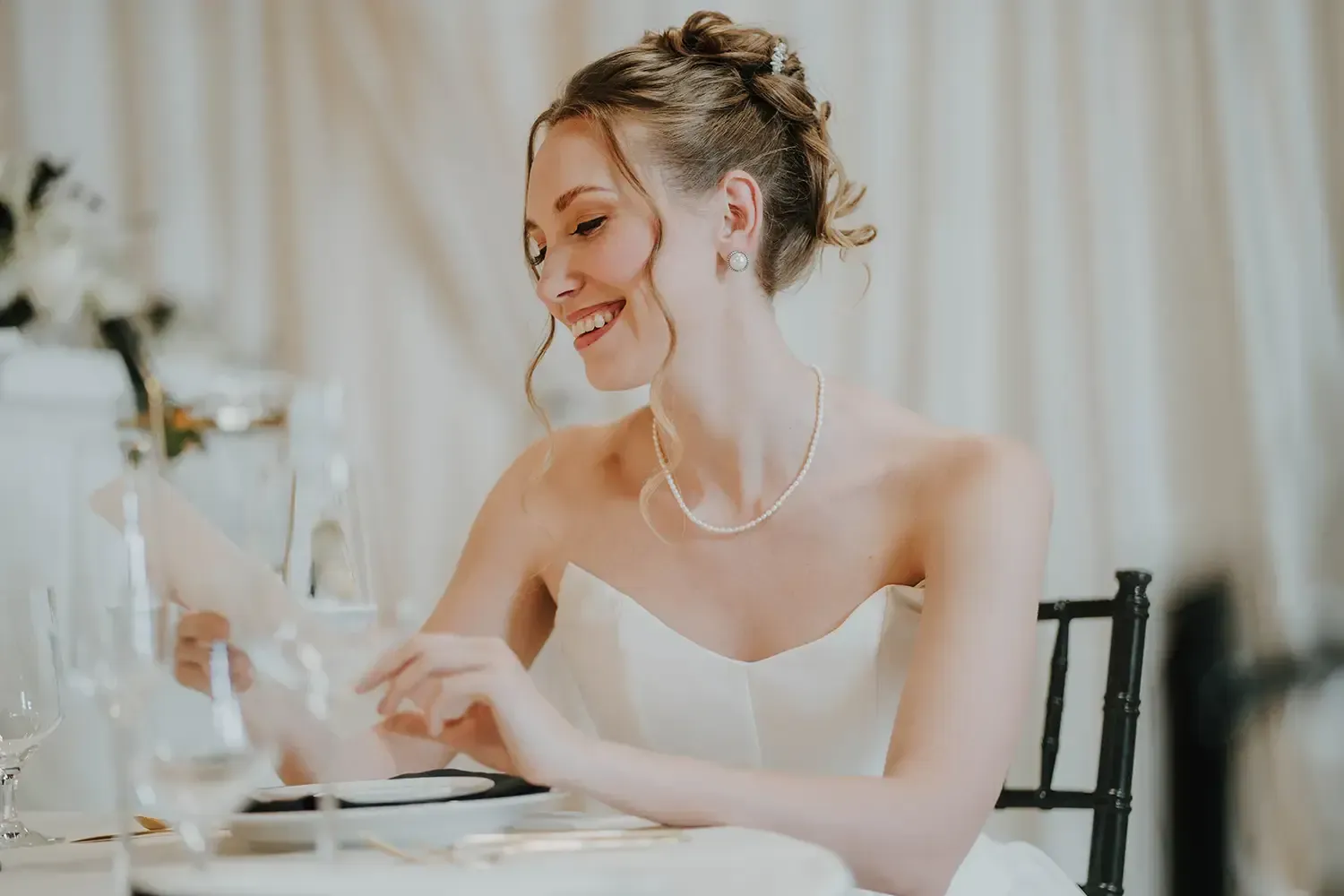 Bride in a white dress smiling at a table, looking at a card