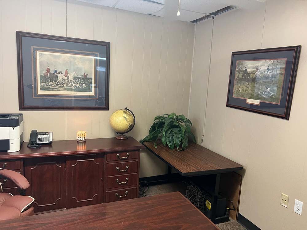 Small office corner with dark wood desk, globe, plants, framed art, and beige walls.