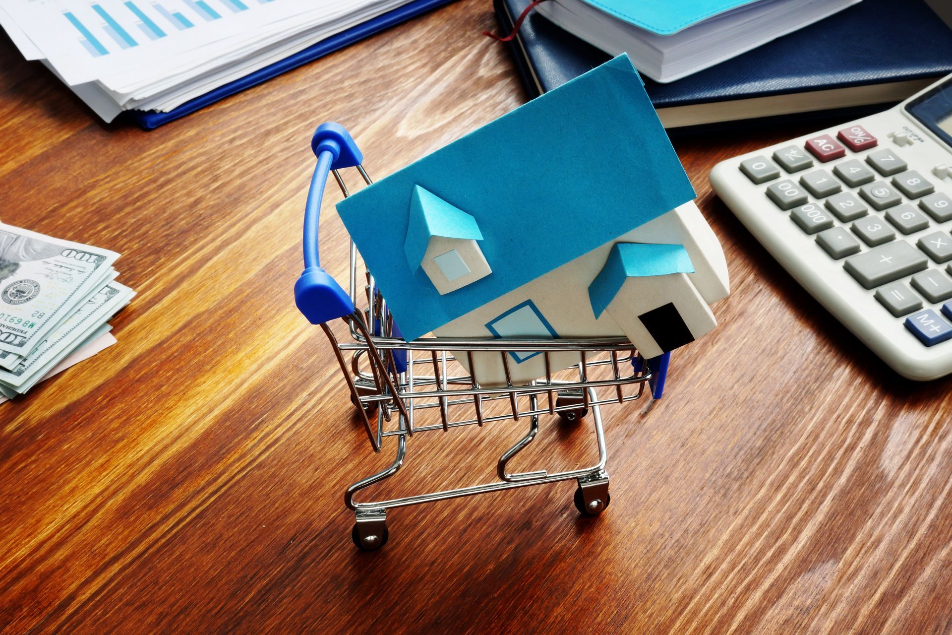Mini shopping cart with blue house model on a desk beside a calculator and paperwork