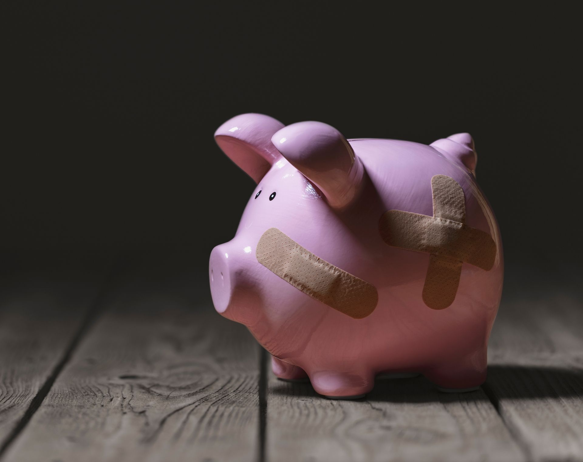 Pink piggy bank with bandages on a wooden floor, lit dramatically against a dark background