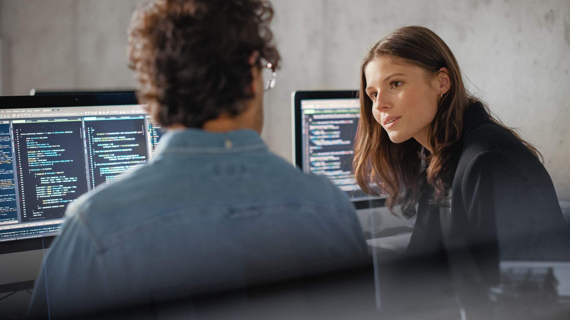 Two people at computer monitors discussing code in a dim office setting