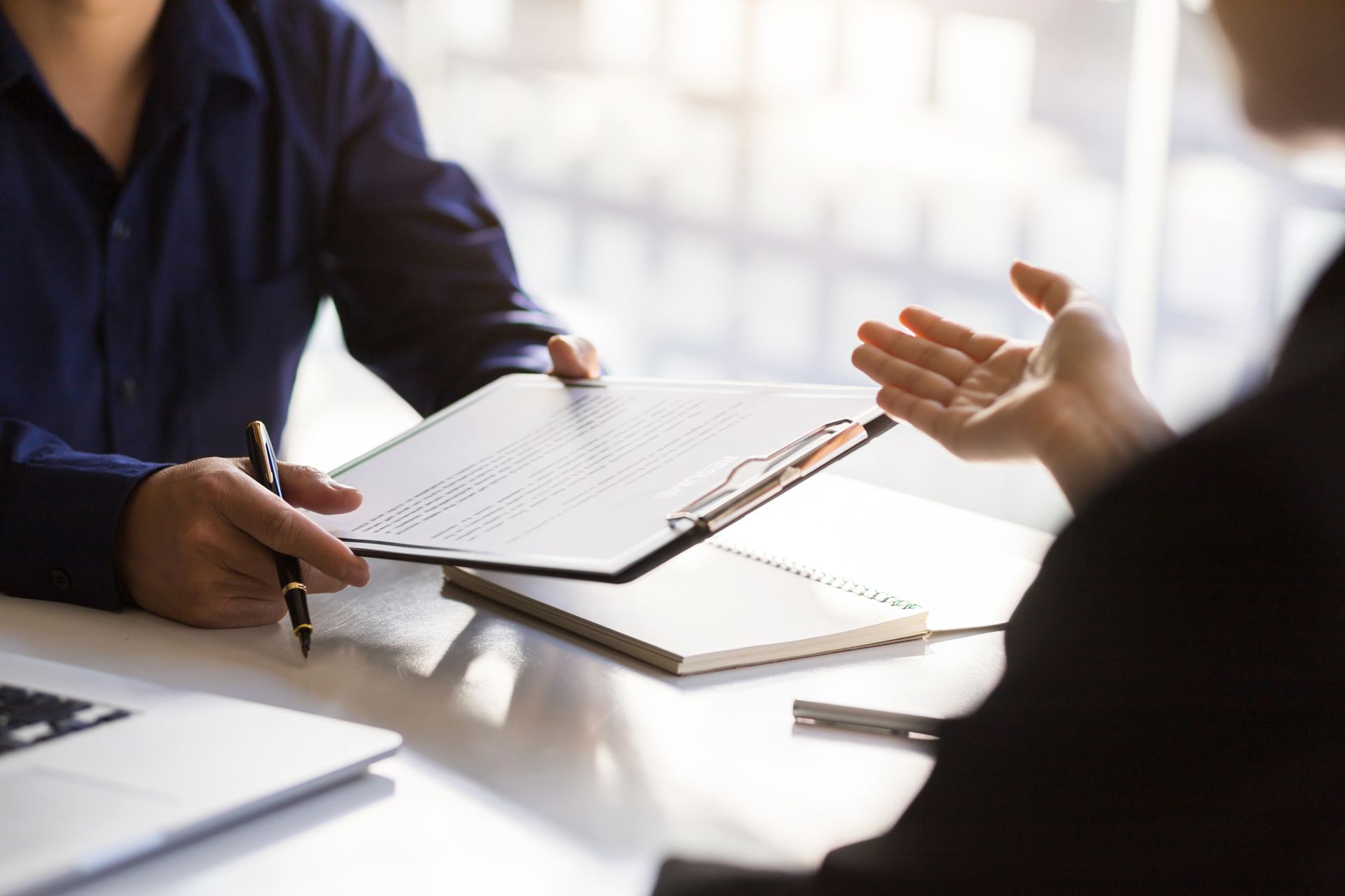 Two people discussing a document at a desk, one taking notes in a bright office