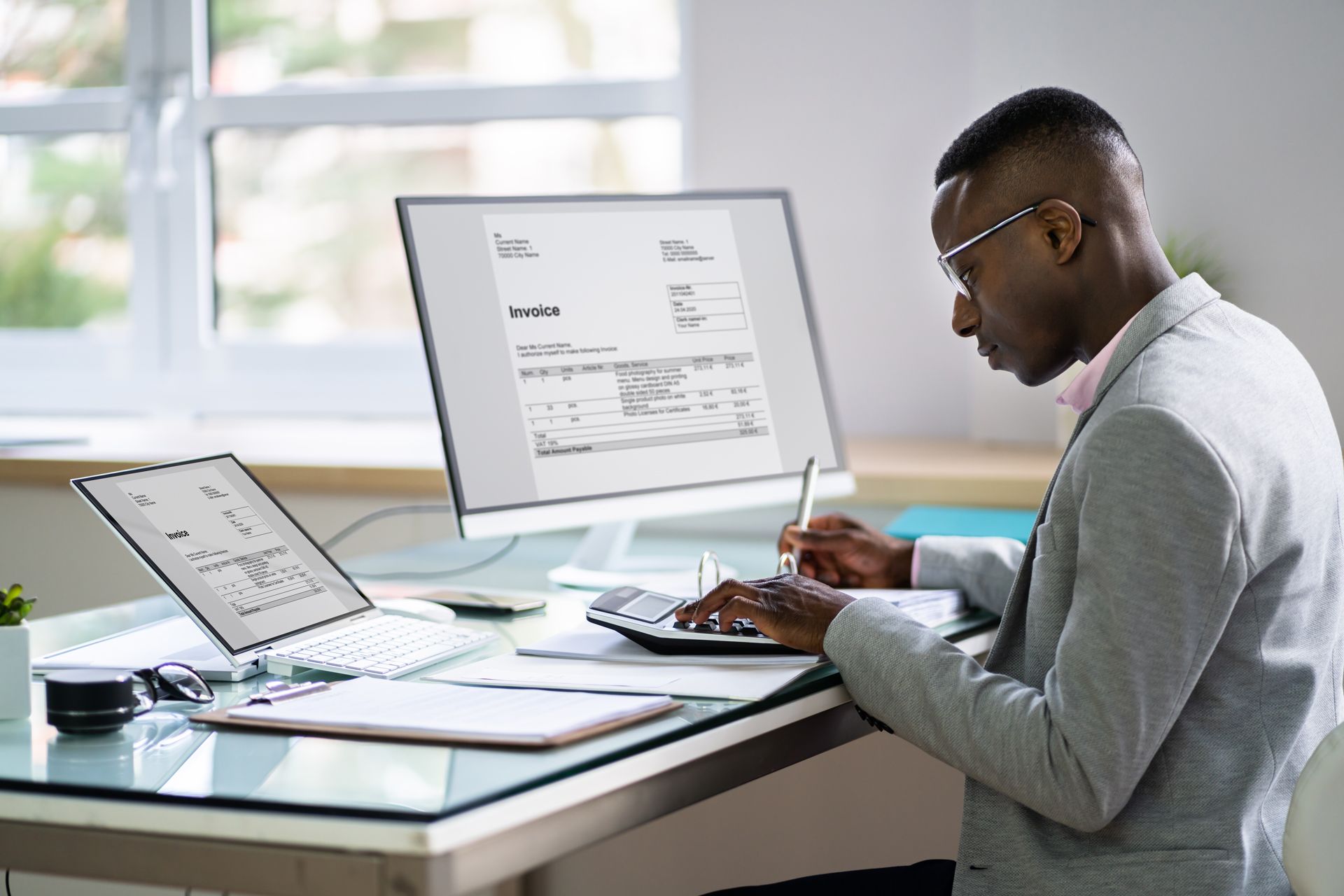 Person working at a desk with multiple screens, writing in a notebook beside a laptop and monitor.