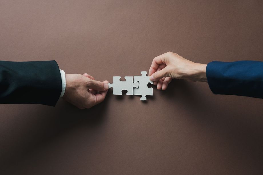 Two hands from opposite sides connect a white puzzle piece on a brown background