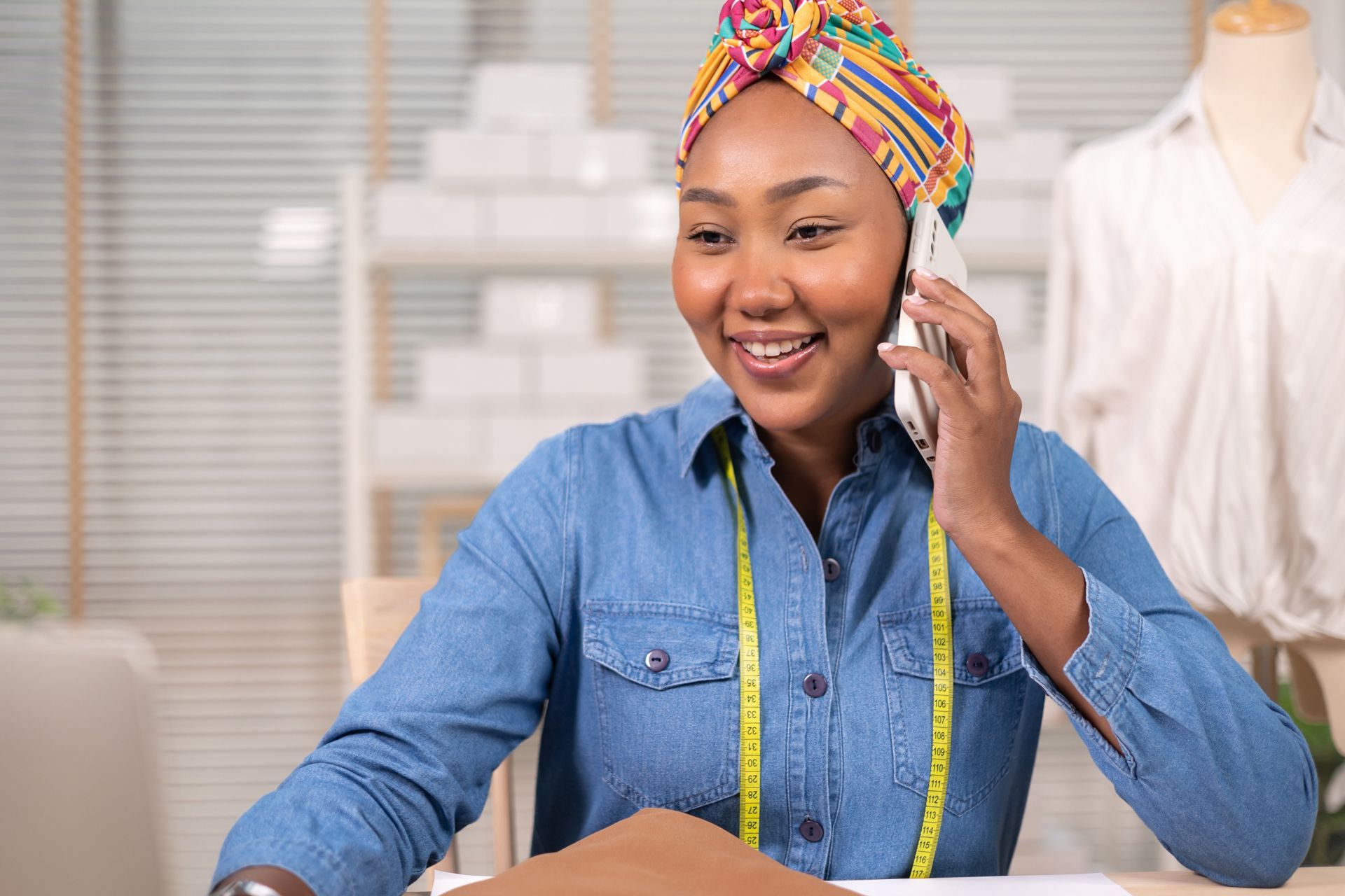 Smiling person in a colorful headwrap talks on a phone in a bright workspace, wearing a denim shirt and measuring tape.