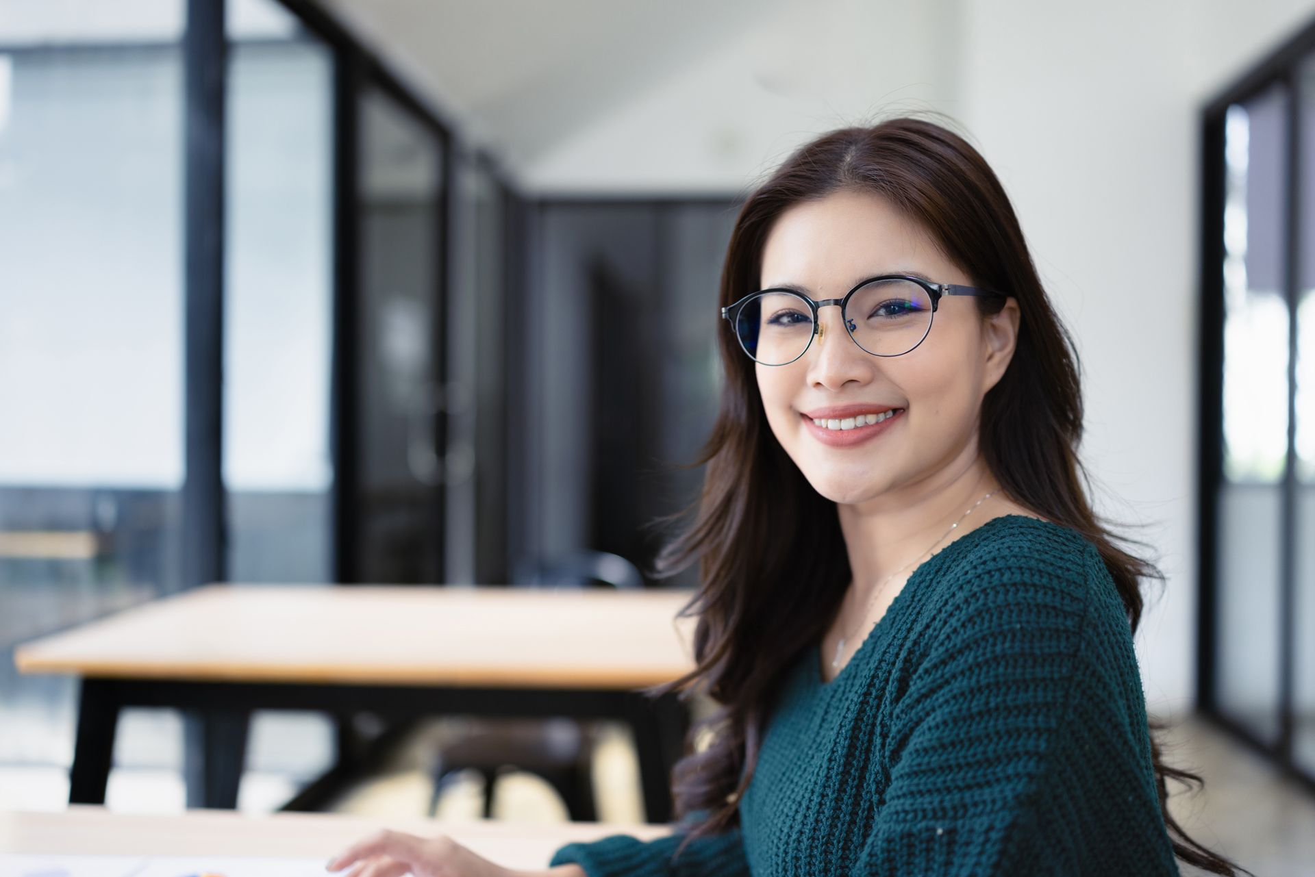 Smiling woman with glasses seated in a bright office or classroom, wearing a teal sweater.