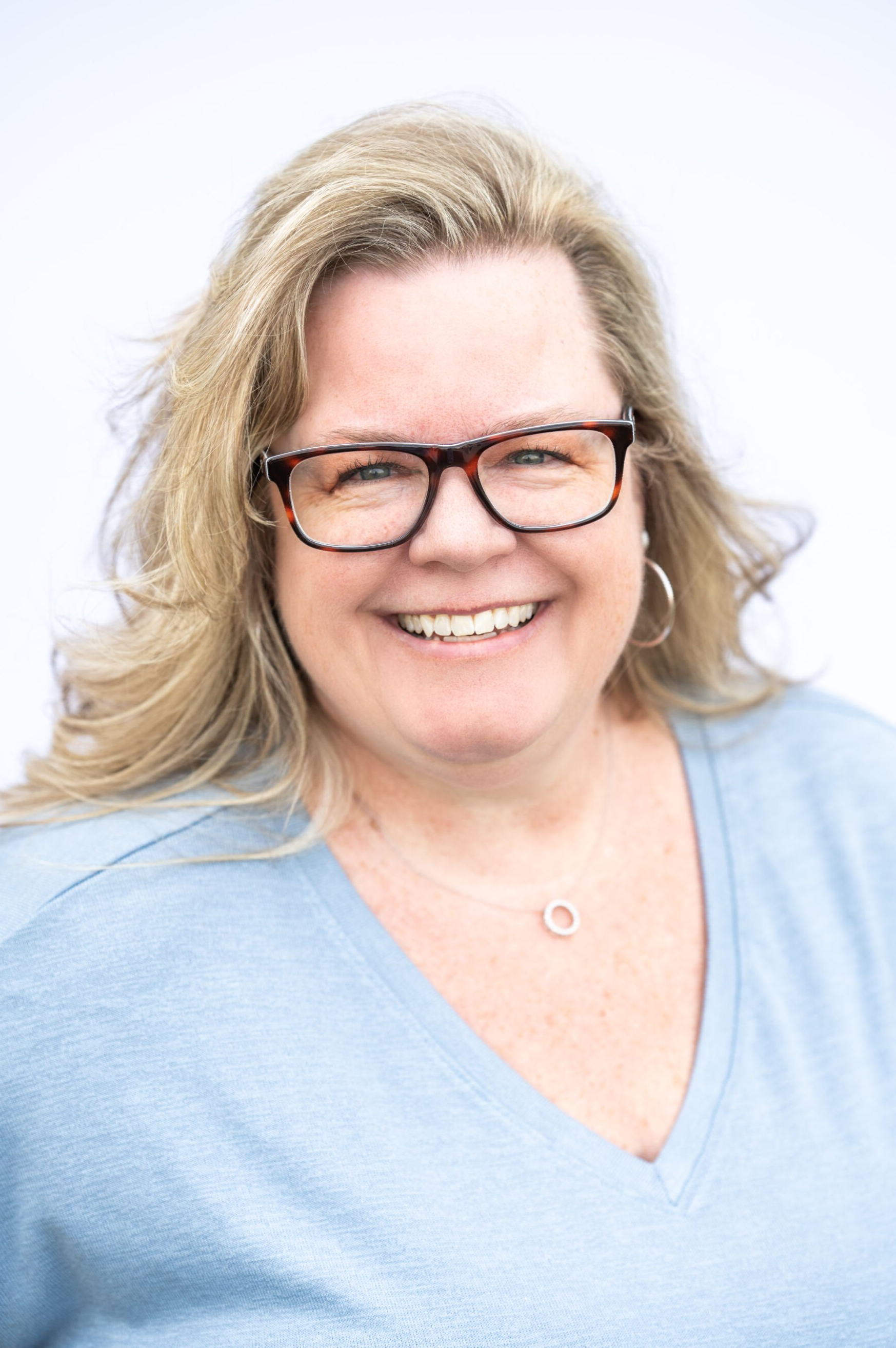 Smiling person with blonde hair and glasses wearing a light blue top against a white background