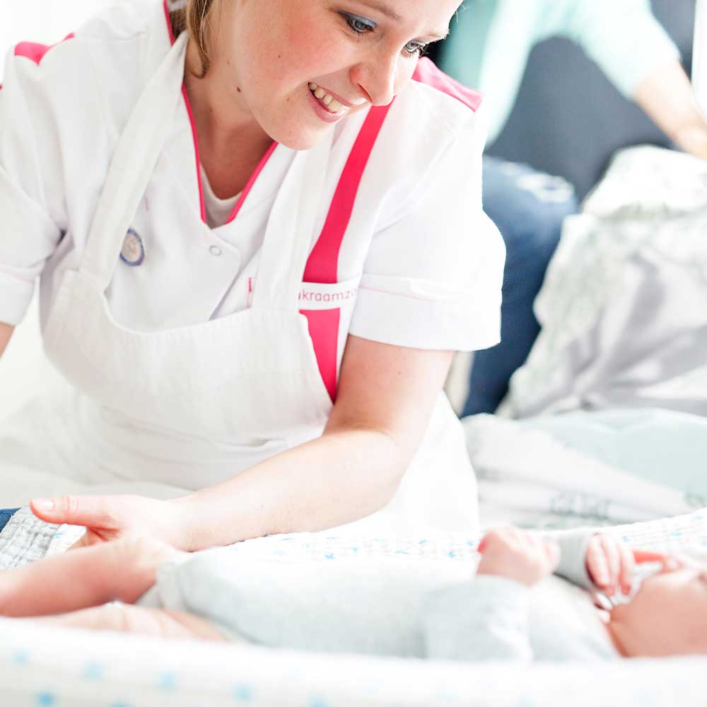 Een glimlachende verpleegster kijkt naar een baby in een wiegje.