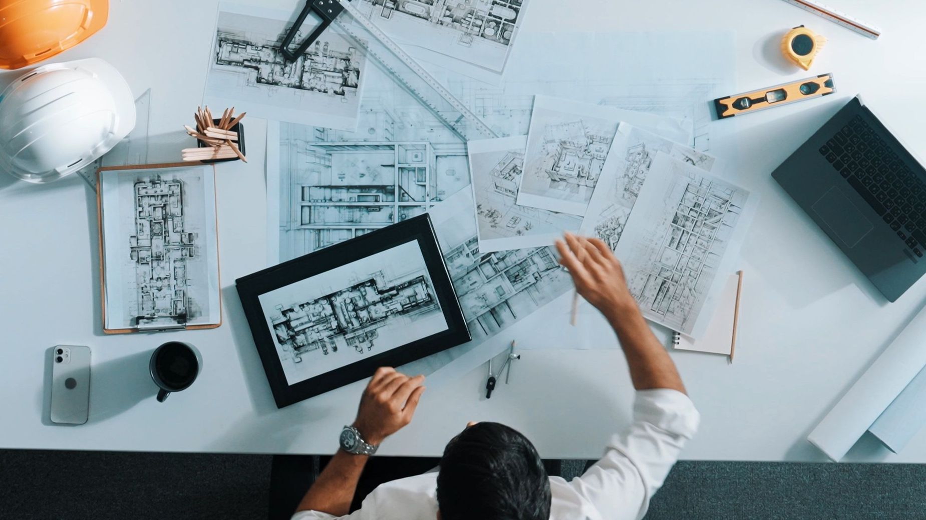 Hände betrachten Architekturpläne, auf einem Schreibtisch stehen ein Tablet, eine Kaffeetasse und ein Laptop.