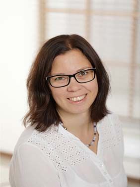 Porträt einer lächelnden Person mit Brille und dunklen Haaren, die eine weiße Bluse trägt, vor einem hellen Hintergrund