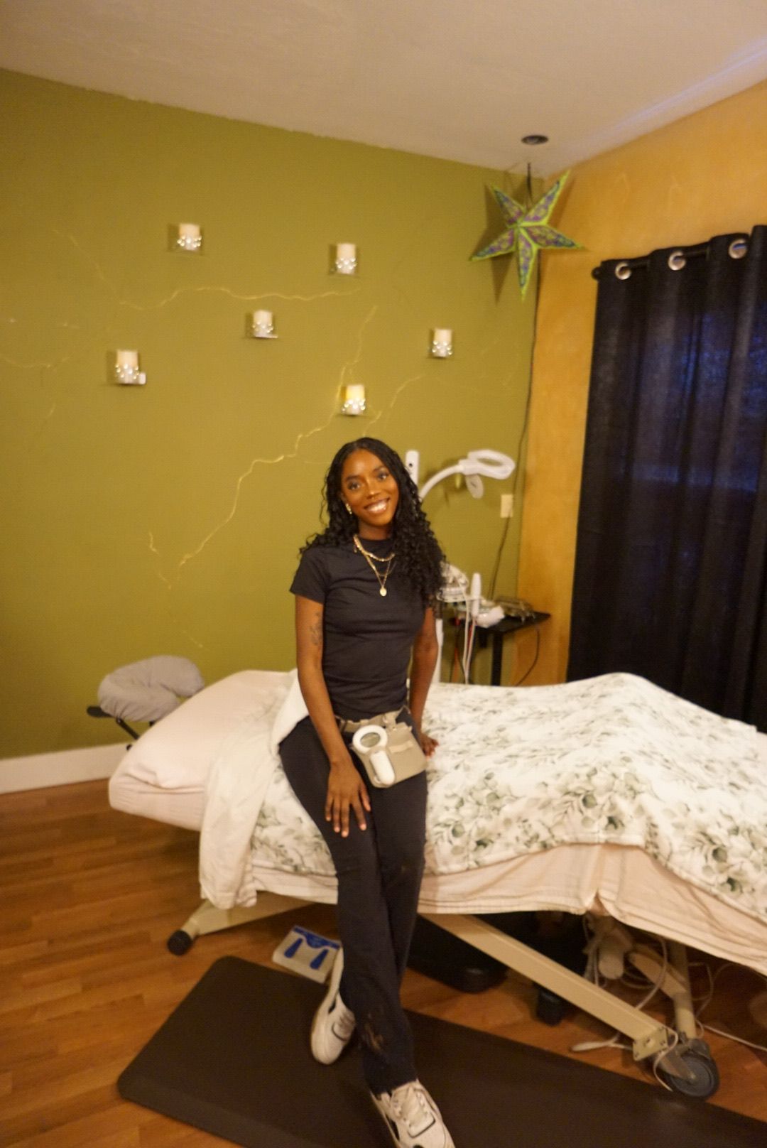 Woman sitting on a bed in a cozy bedroom with green walls, black curtains, and soft lighting.