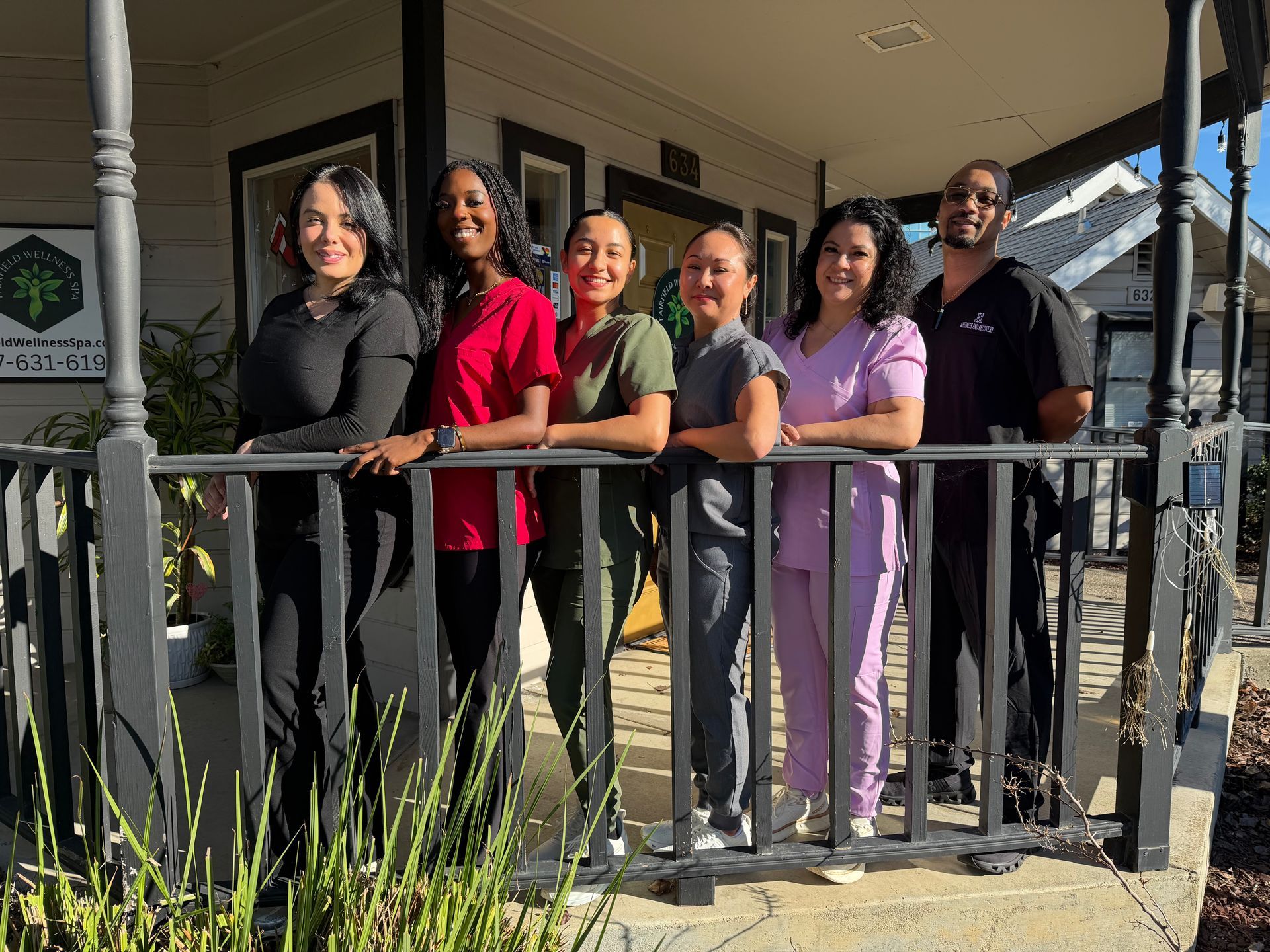 Five people standing on a porch, smiling behind a black railing in bright sunlight.