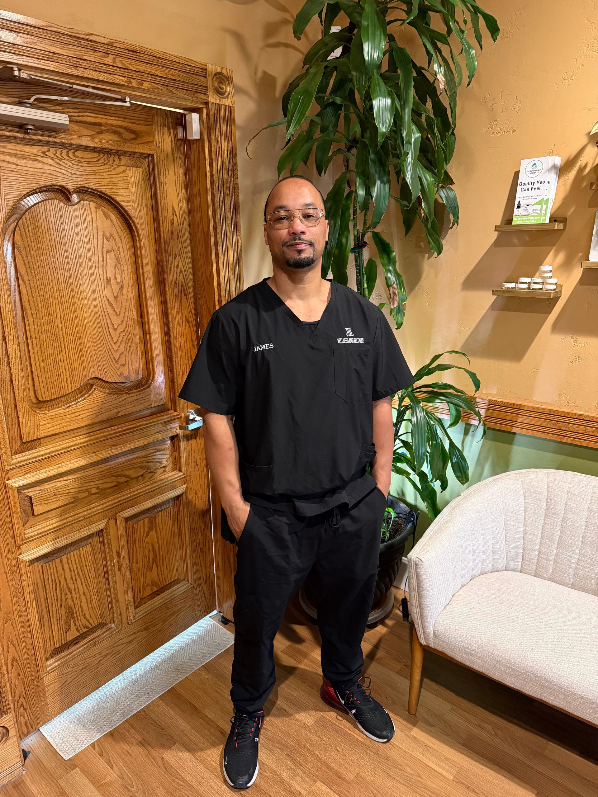 Person in black scrubs standing in a warmly lit room with carved wooden doors and a large plant.