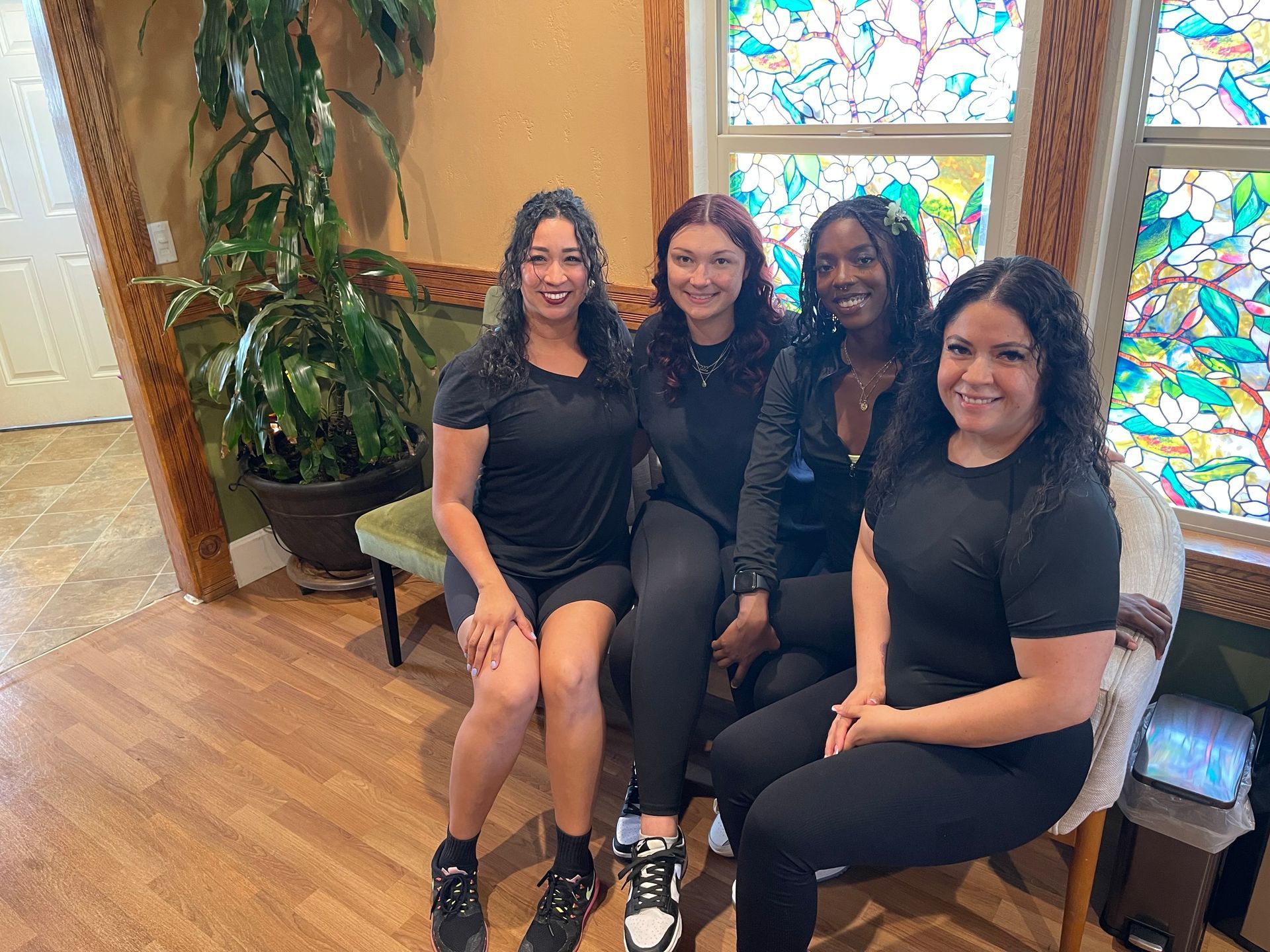 Four people in black outfits sitting together on a bench in a bright room with stained glass windows.