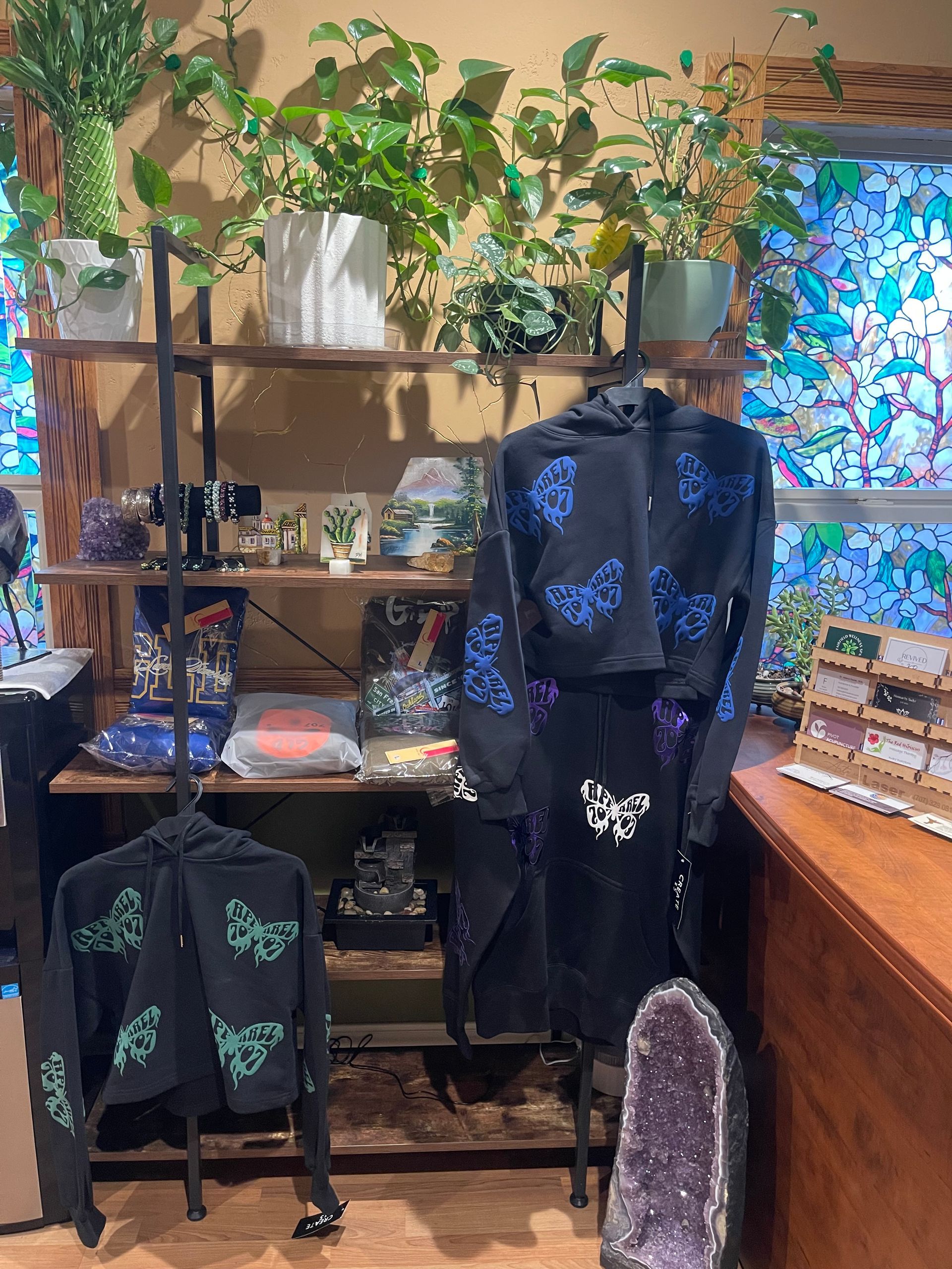Black-and-green floral clothing displayed on racks and shelves in a cozy boutique with hanging plants.