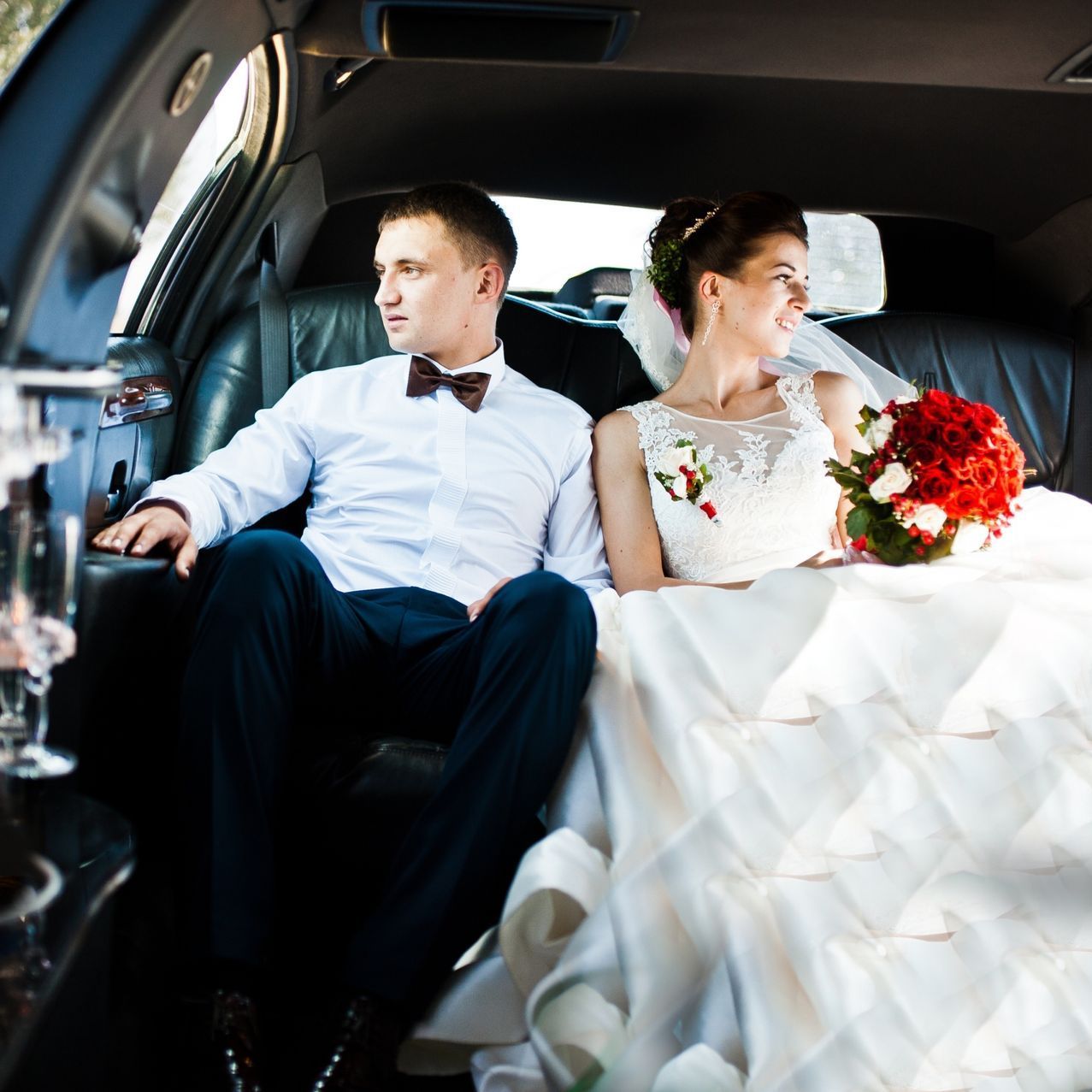 Bride and groom seated in a limousine, bride holding a red bouquet, both looking away from each other