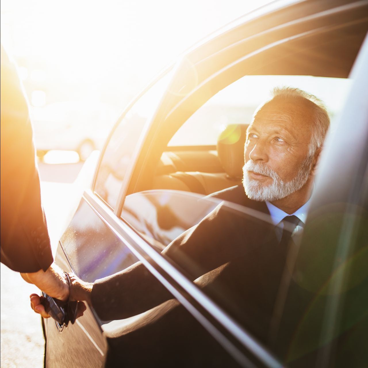Man in a suit shaking hands with someone through a car window in warm sunlight