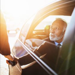 Man in a suit shaking hands with someone through a car window in warm sunlight