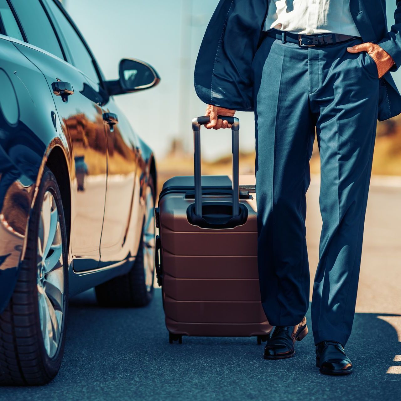 Traveler in navy pants pulls a brown suitcase beside a parked car on a sunny road.