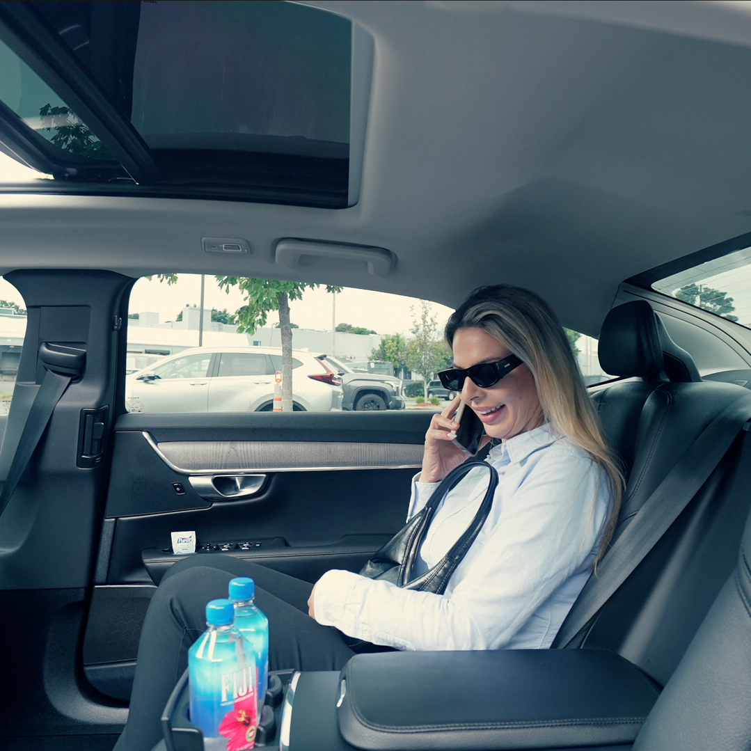 Woman in sunglasses sitting in a car, talking on a phone with a water bottle in the center console.