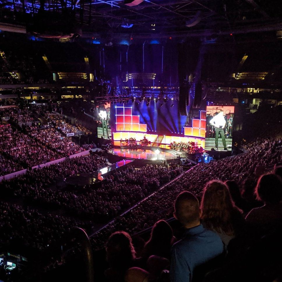 Concert arena packed with crowd, brightly lit stage, and large screens during a live performance