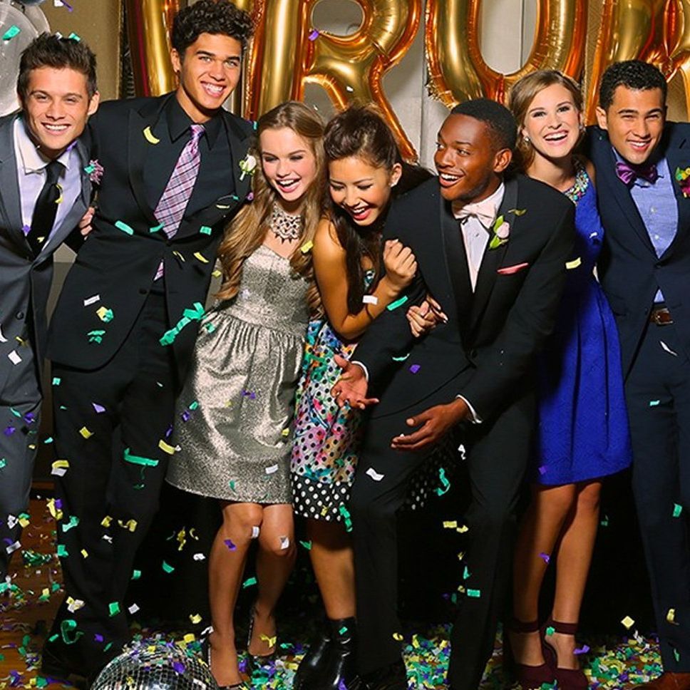 Group of six people posing and laughing at a confetti-filled party, with gold balloons in the background