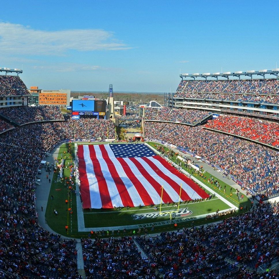 A packed football stadium with a giant American flag stretched across the field during a ceremony.