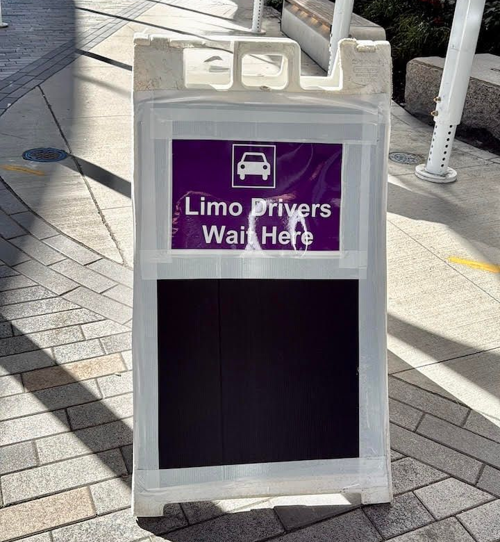 White sidewalk sign with purple panel reading “Limo Drivers Wait Here” and a car icon