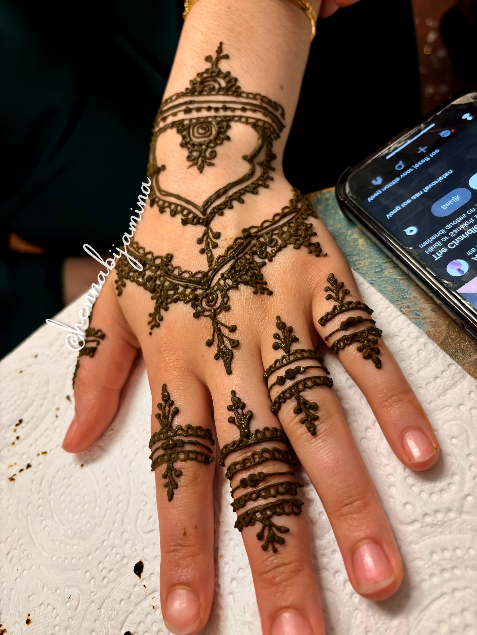 Hand met ingewikkeld henna-ontwerp in bruin op een wit oppervlak, naast een smartphone.