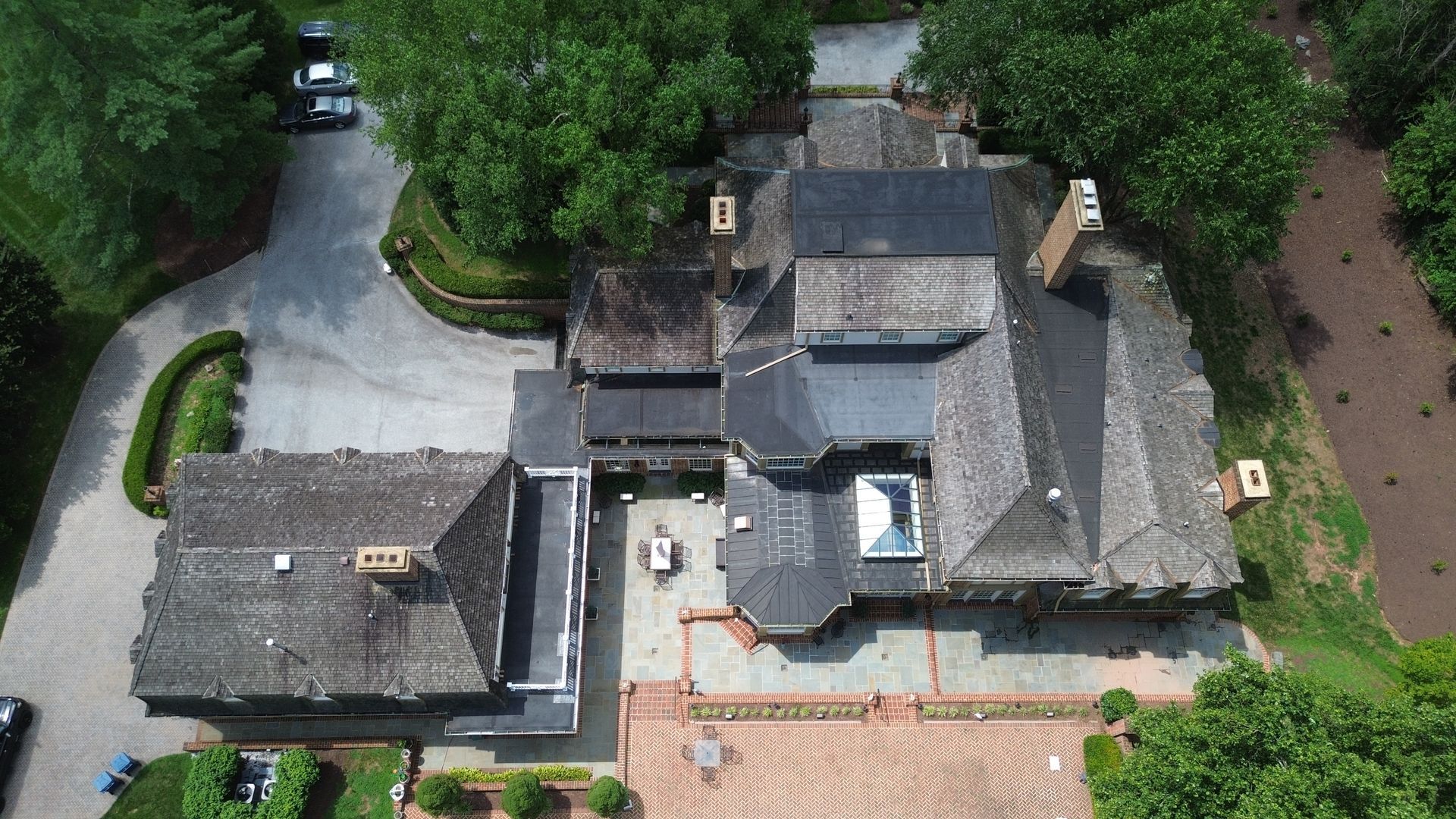 Aerial view of a large gray-roofed house with a driveway, patio, and surrounding trees.