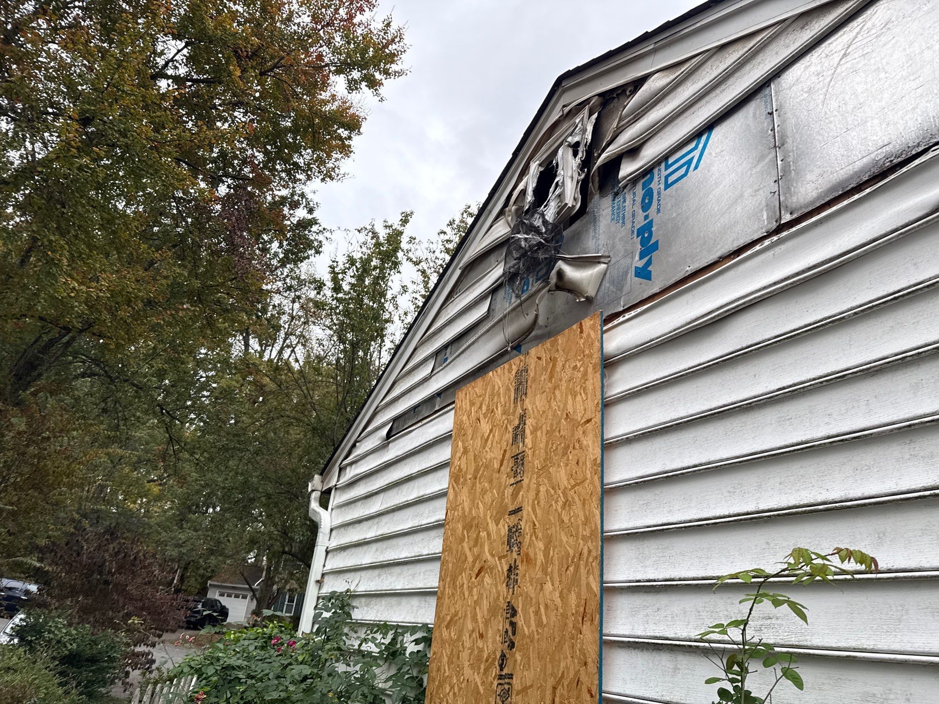 Boarded-up side of a white house with damaged roof trim and a boarded window opening.