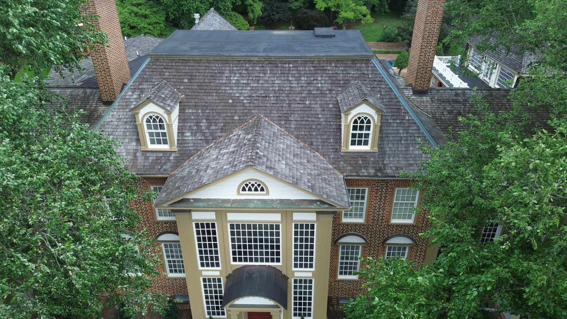 Aerial view of a brick house with a shingled roof, dormer windows, and white columns, surrounded by trees