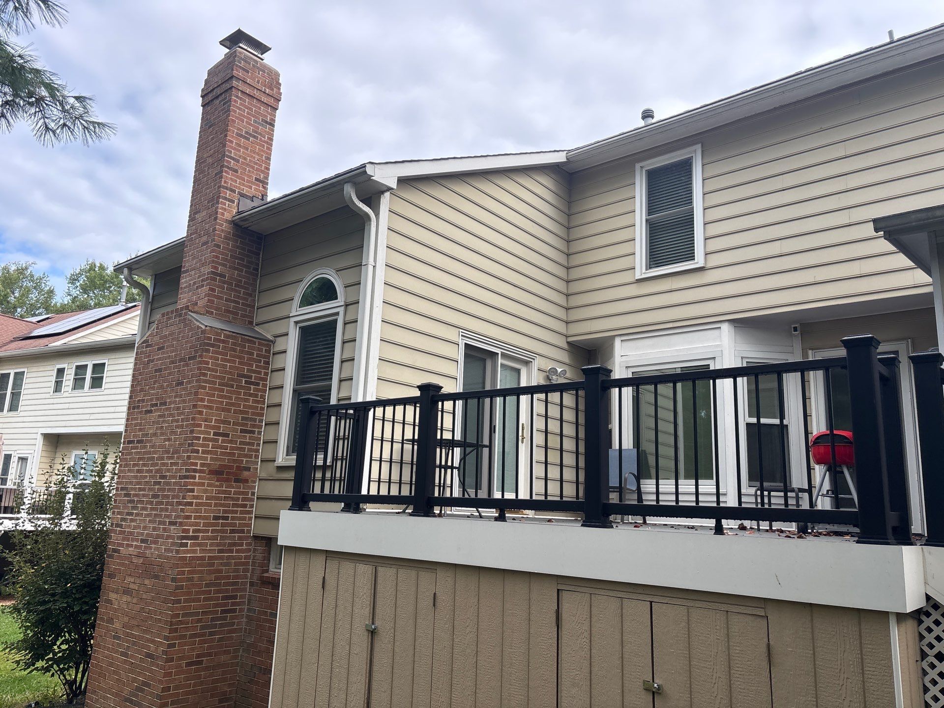 Suburban house exterior with beige siding, brick chimney, black railing, and raised deck