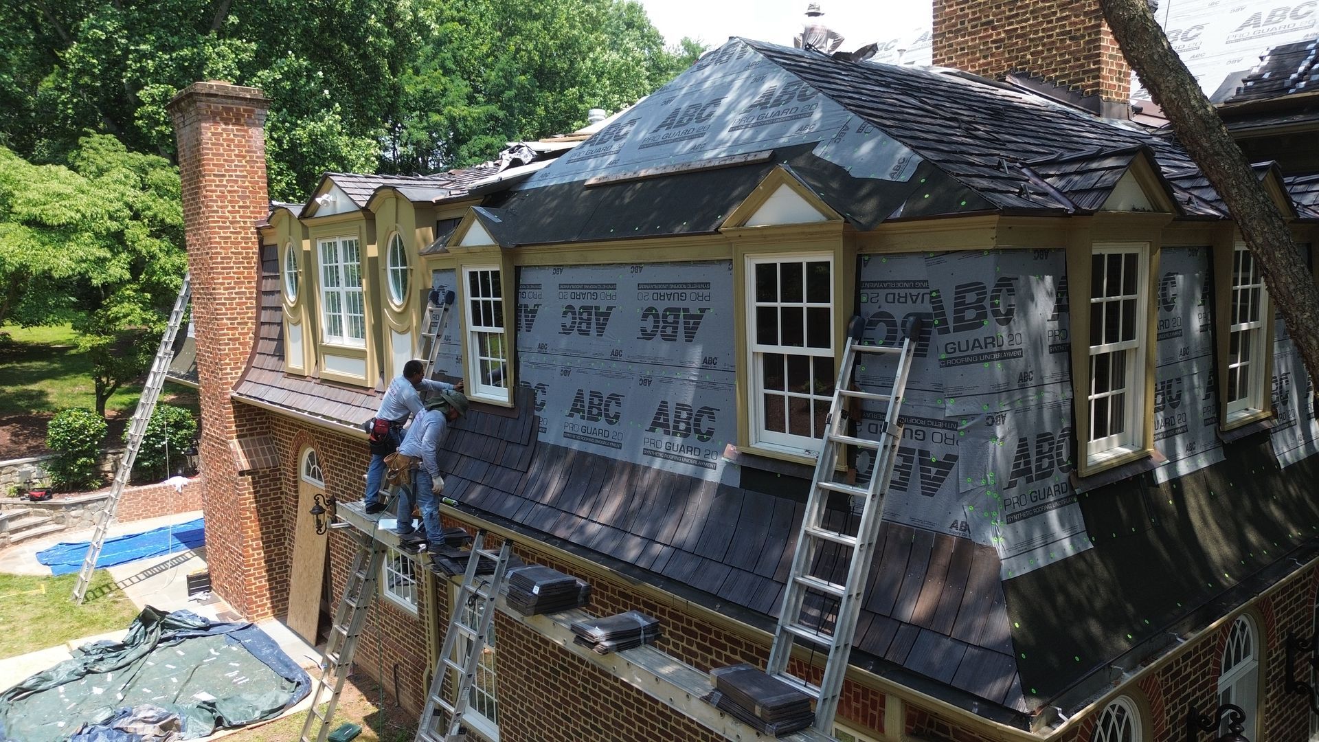 House under renovation with gray tarp covering the roof and workers on a brick chimney