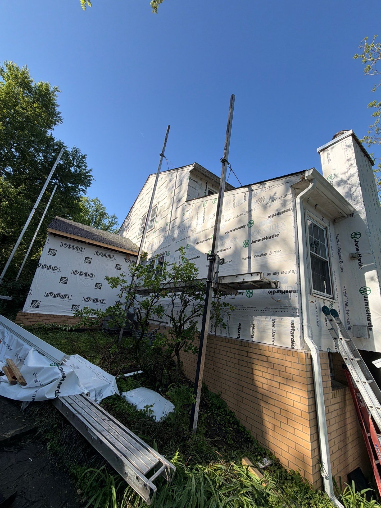 Damaged white house exterior with scattered debris and bent metal under trees in daylight