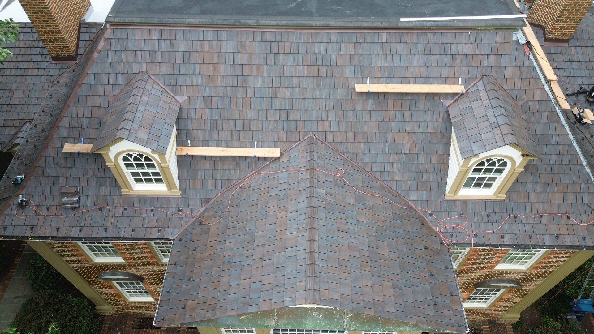 Aerial view of a house roof with gray shingles, dormer windows, and two wooden roof planks.