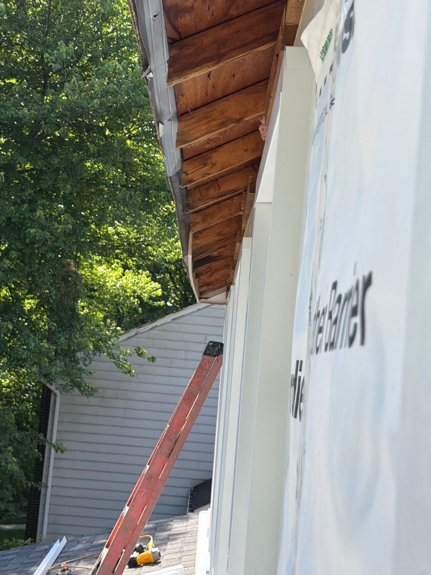 House exterior with white siding, overhanging roof, red ladder, and tree shade during repairs