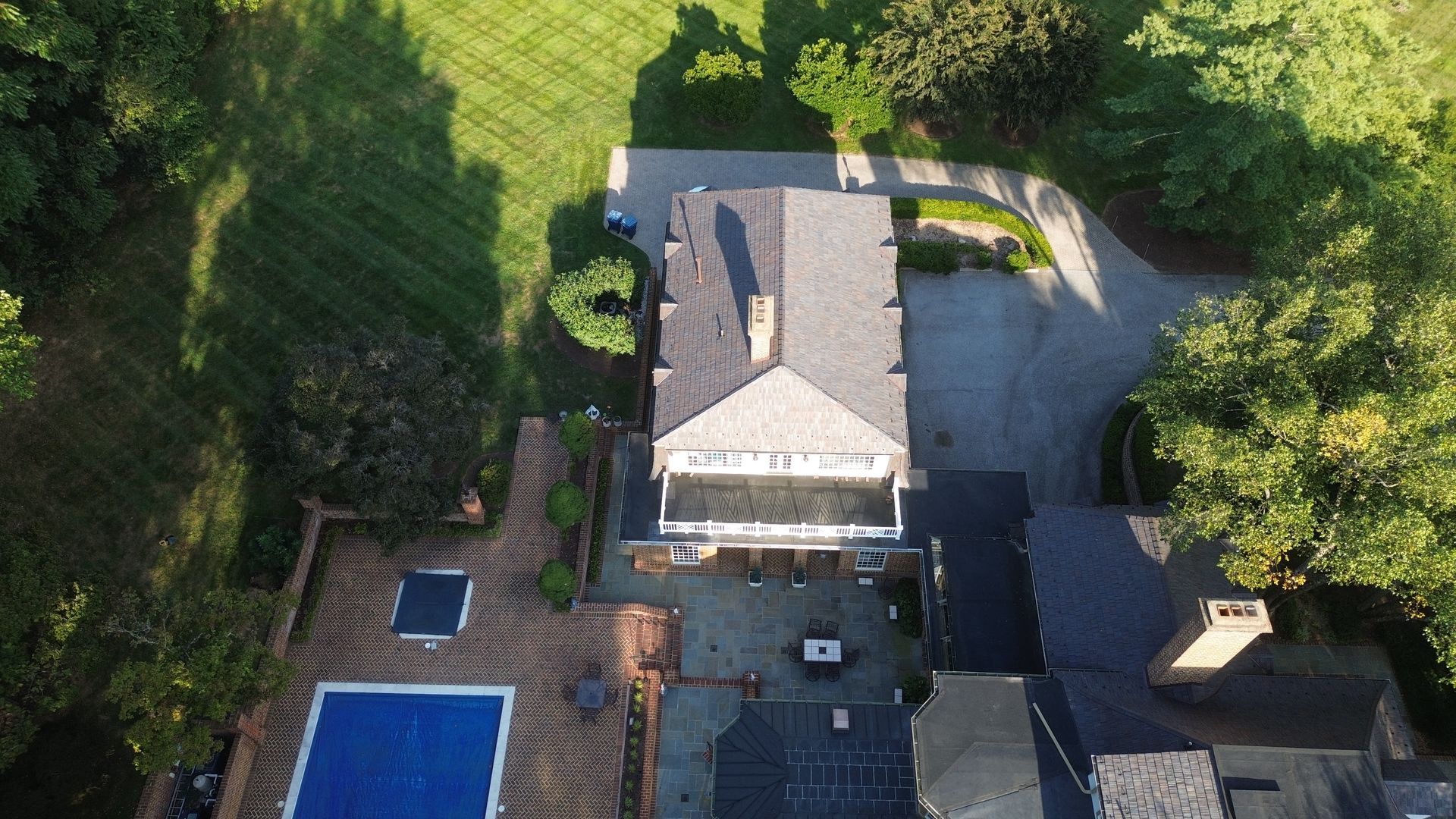 Aerial view of a house with driveway, pool, patio, and surrounding trees and lawn.