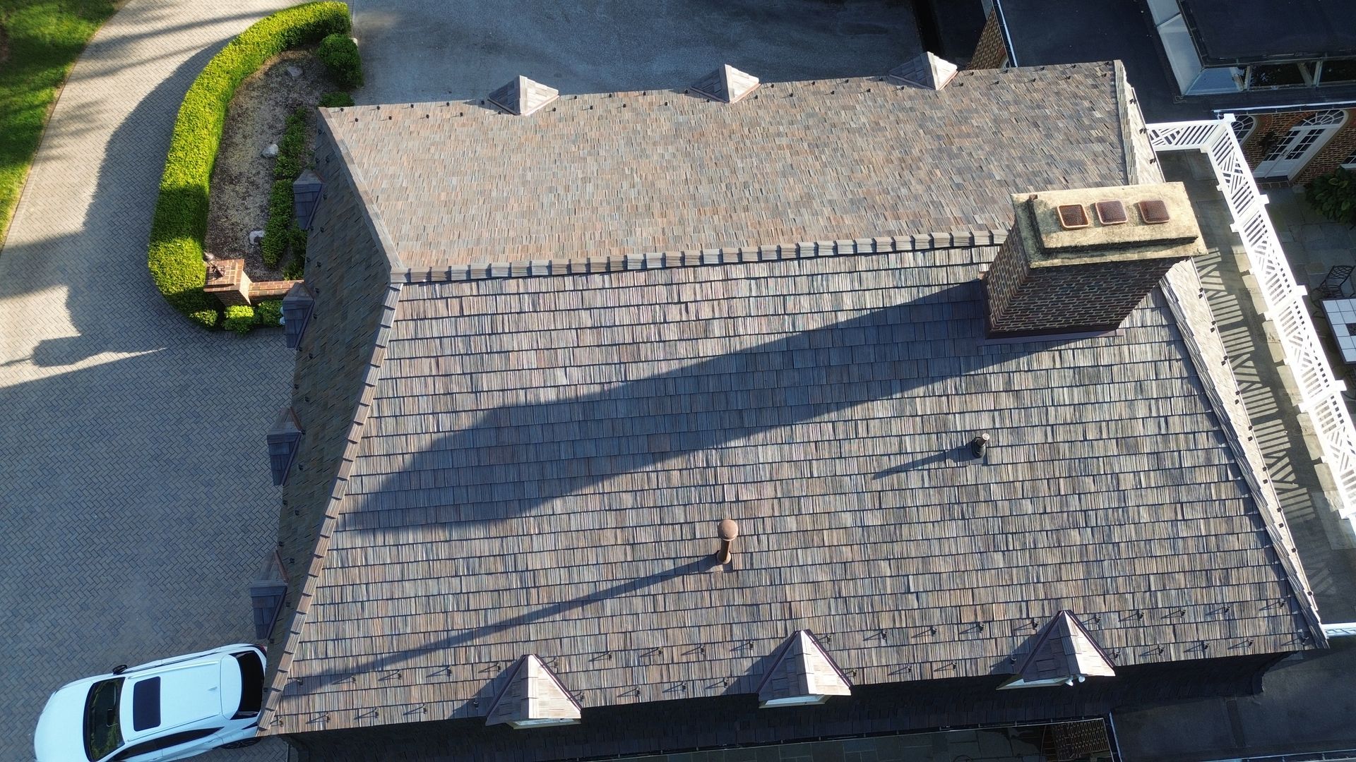 Aerial view of a gray shingled house roof with a chimney and a parked car at the edge