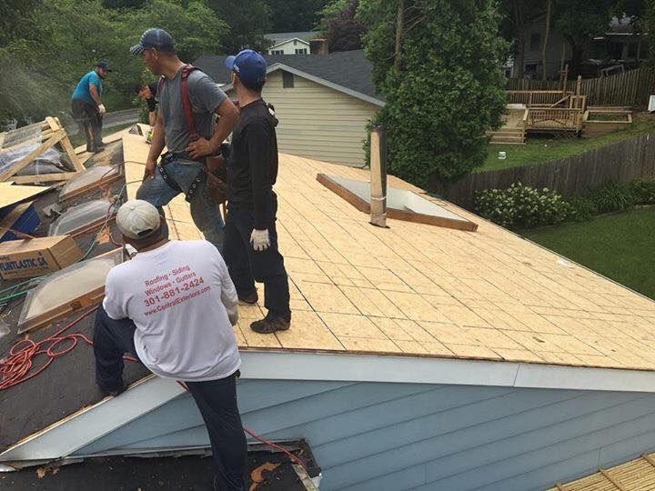 Four construction workers wearing safety gear work on a sloped residential roof with exposed plywood decking.
