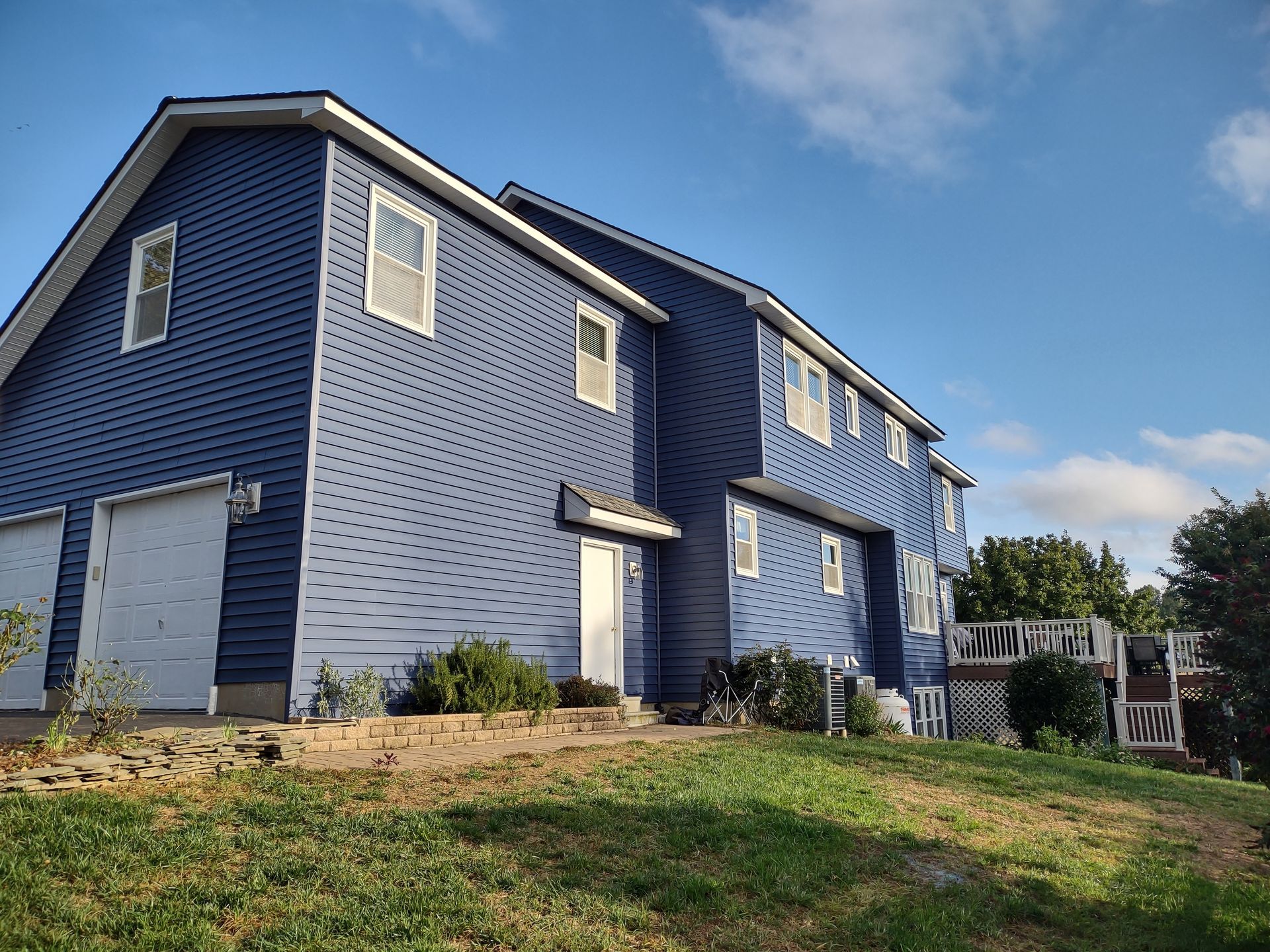 Blue two-story house with white trim on a grassy slope under a partly cloudy sky