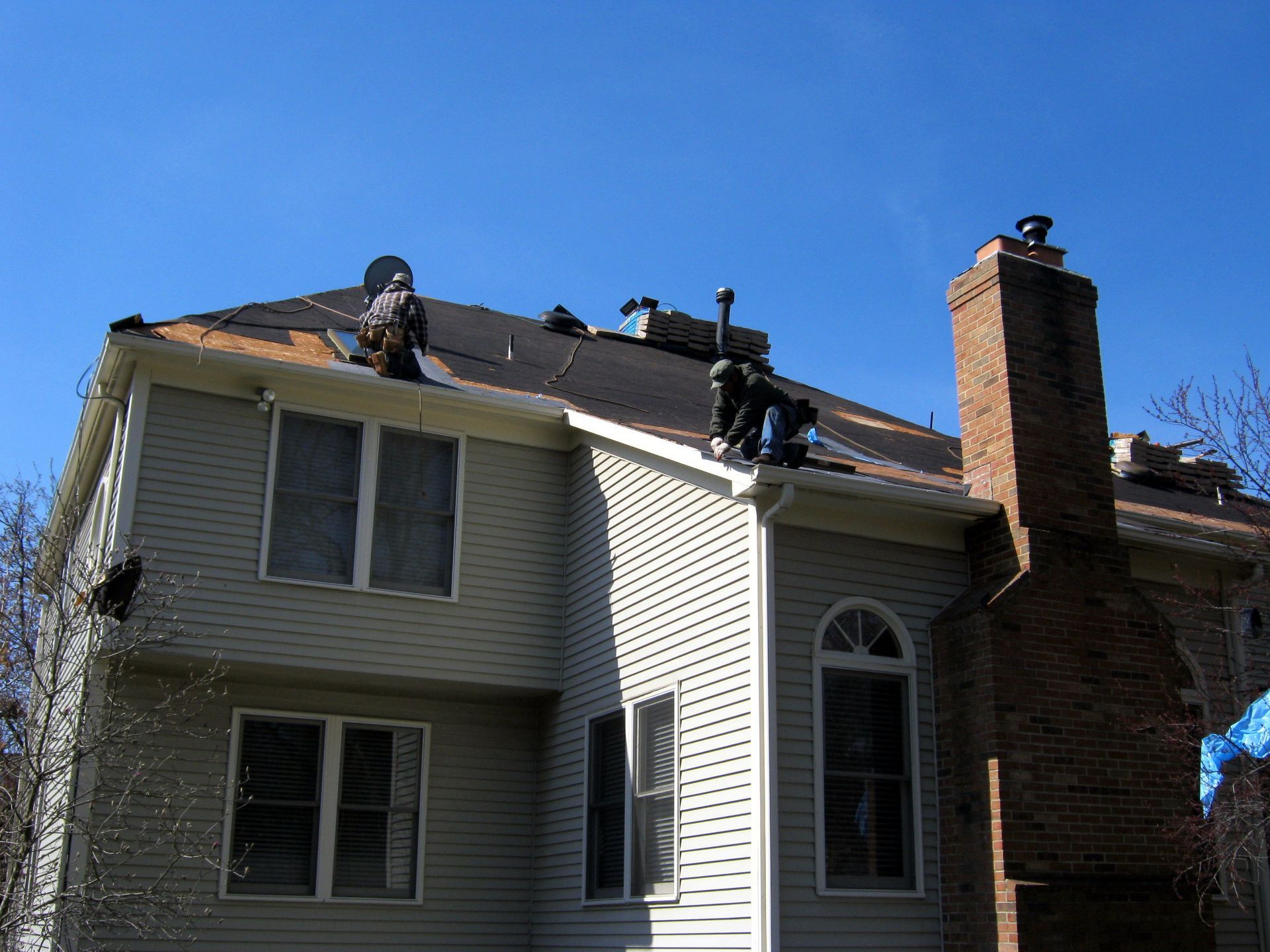 Workers repairing a roof on a two-story house with a brick chimney under a clear blue sky