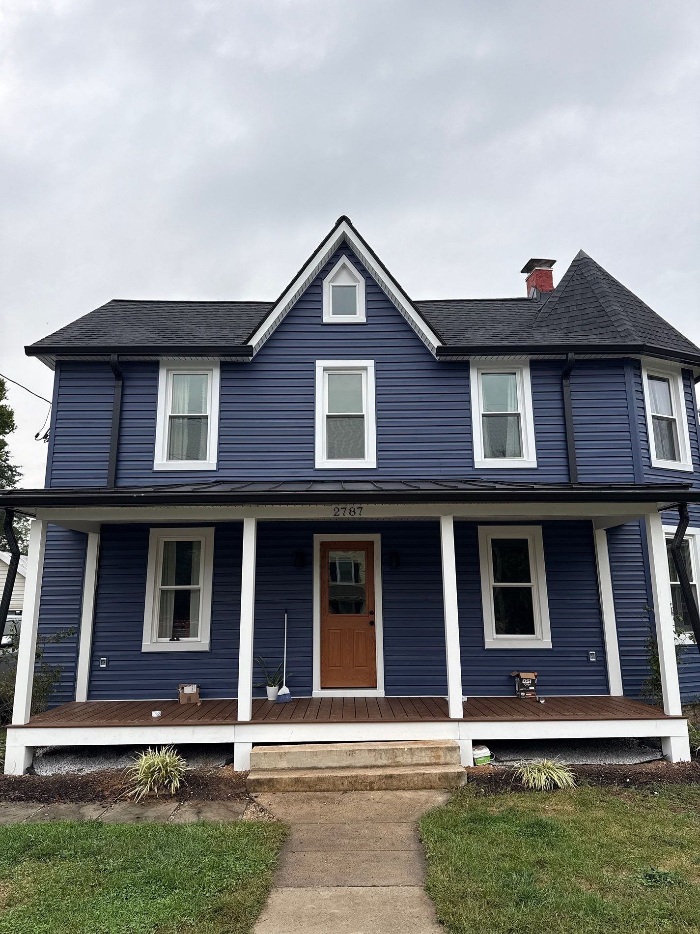 Two-story blue house with white trim, brown door, and covered porch under an overcast sky.
