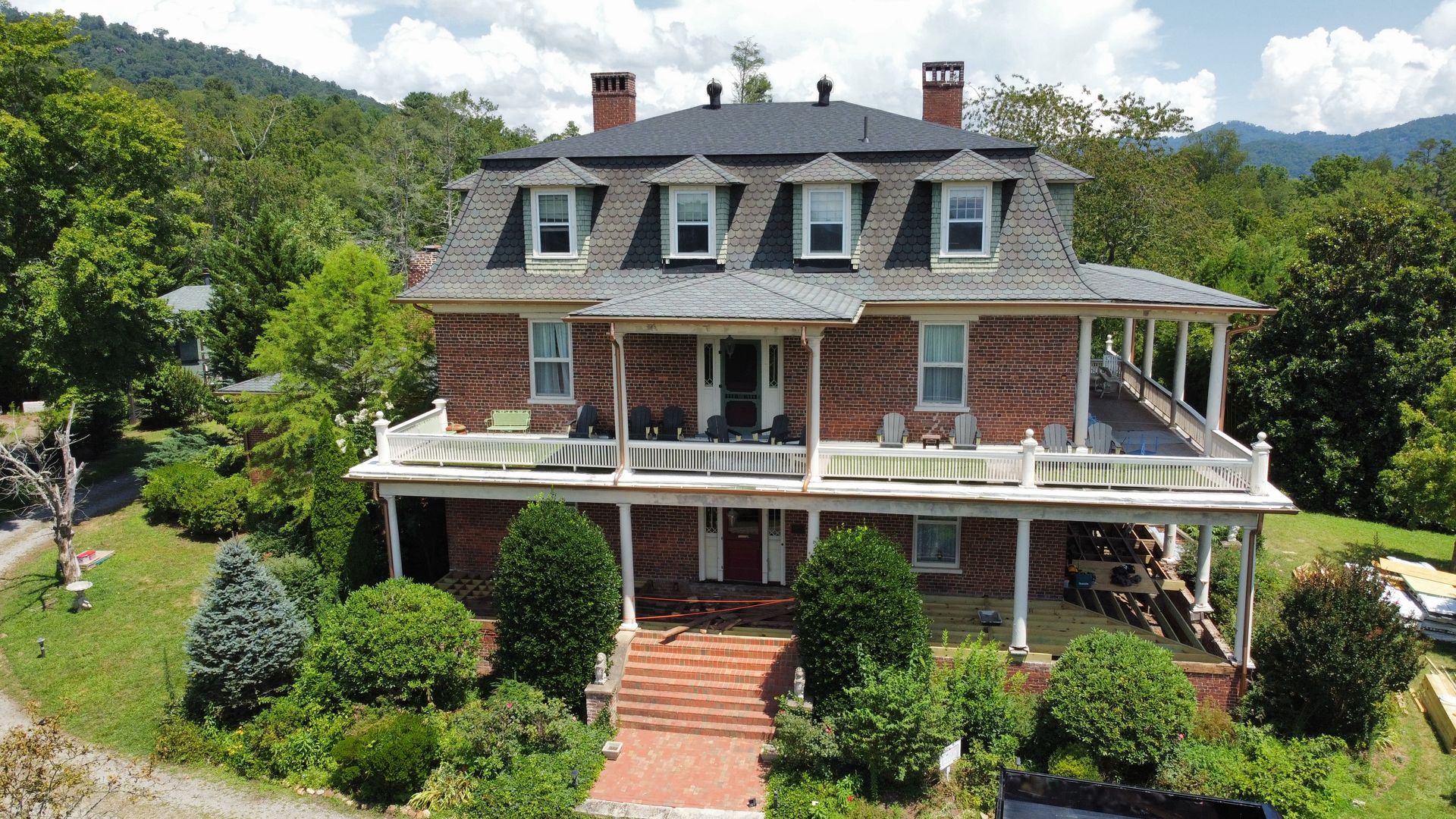Large brick house with a wraparound porch, dormer windows, and a landscaped front yard in a mountain setting