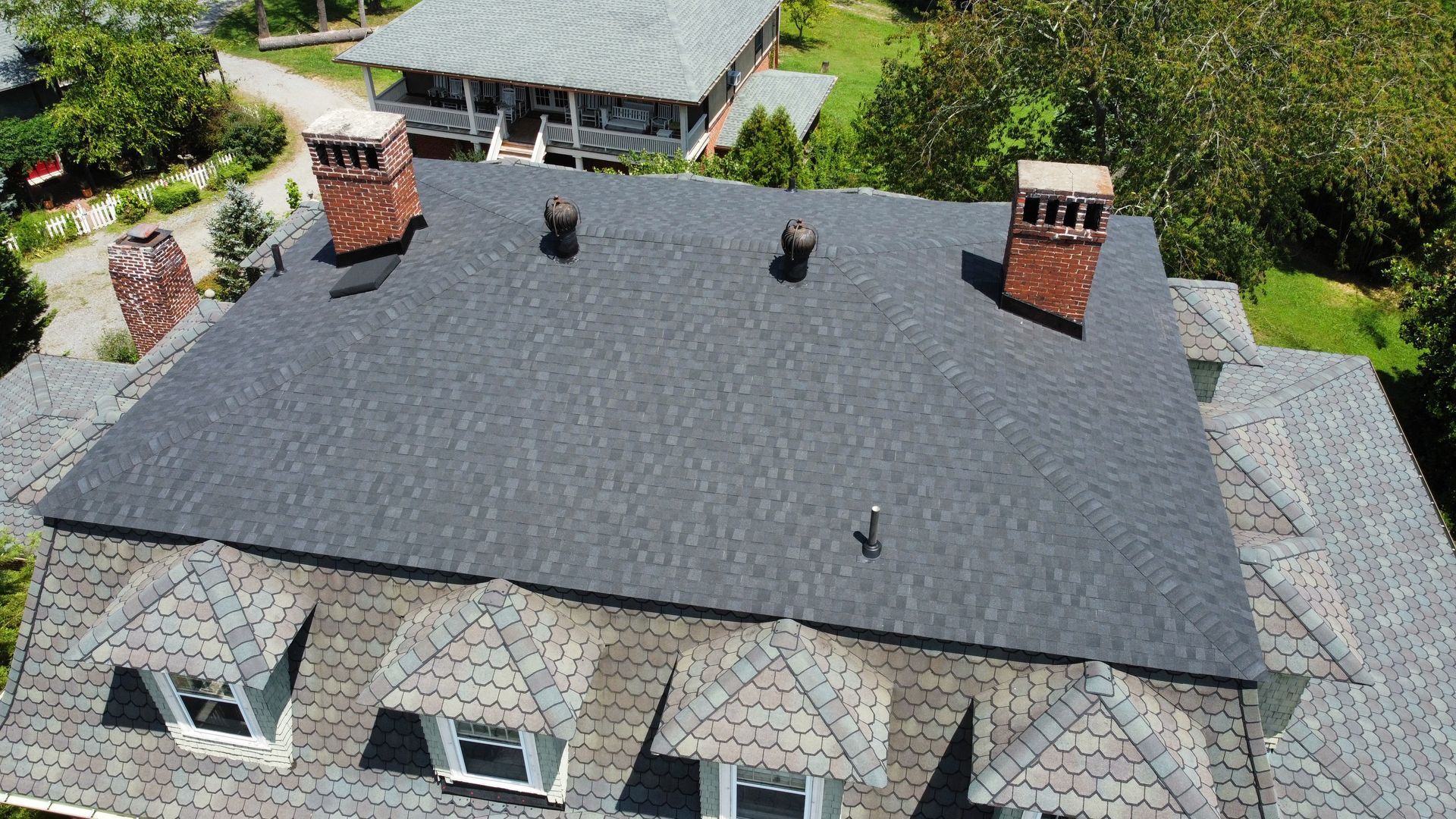 Aerial view of a house with a dark gray roof, two brick chimneys, and skylights amid green trees