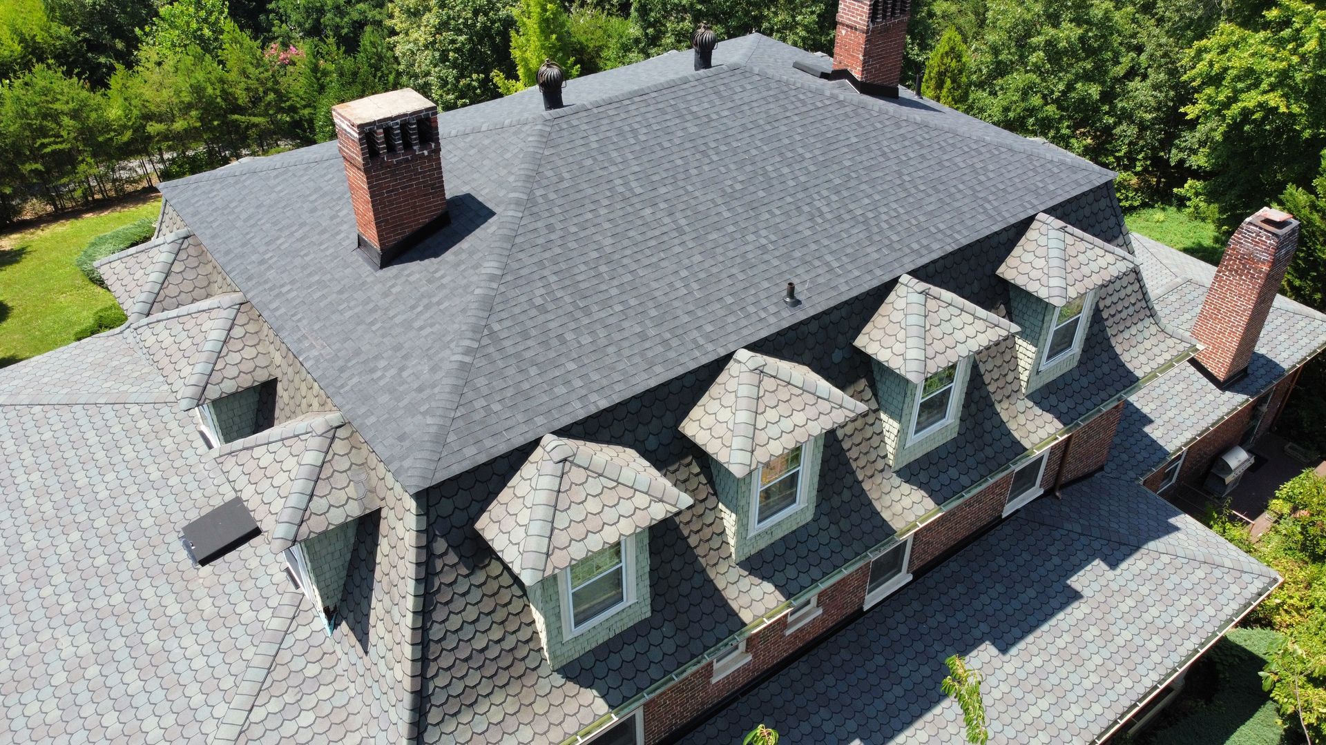 Aerial view of a gray shingled house roof with dormer windows and red brick chimneys surrounded by trees