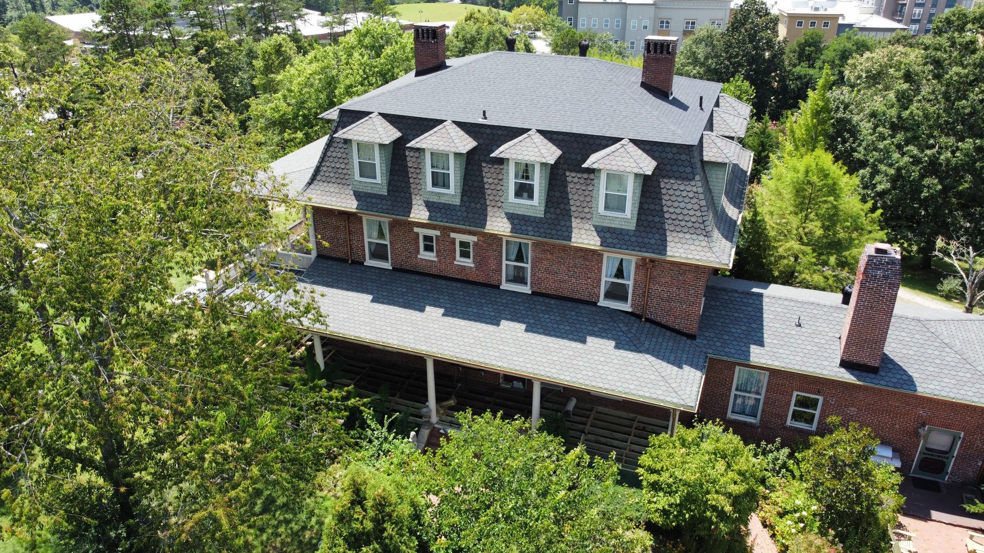 Aerial view of a large brick house with gray roof and dormer windows, surrounded by dense green trees.