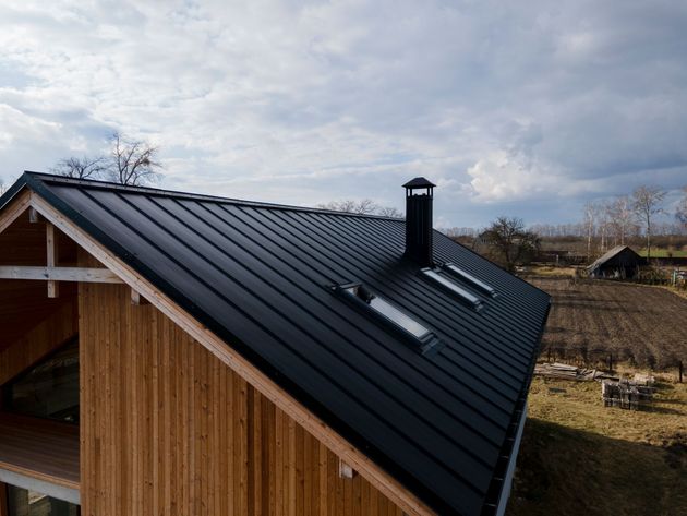 Black metal roof with skylights and chimney on a wooden house under a cloudy sky
